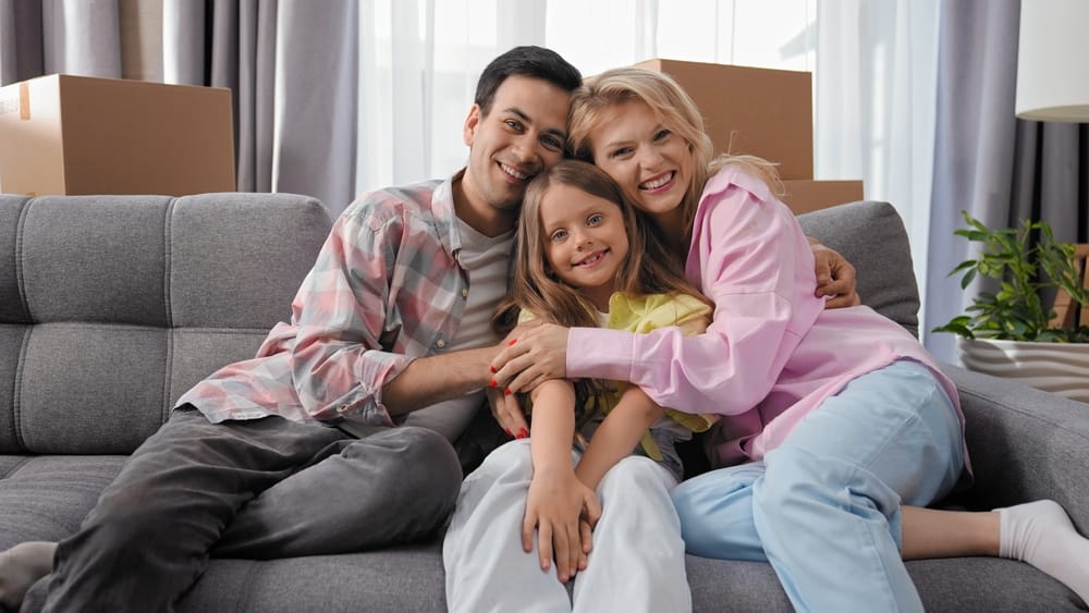 A cheerful family of four sits together on a rug in their living room, with the parents forming a roof shape with their arms over their children - Almost Home