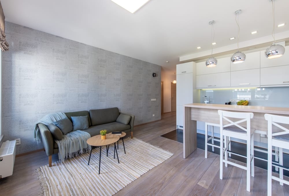 A modern, open-concept living room and kitchen with a gray sofa, a small round table, and a kitchen island with two bar stools under hanging lights - Almost Home A modern, open-concept living room and kitchen with a gray sofa, a small round table, and a kitchen island with two bar stools under hanging lights - Almost Home